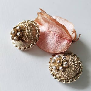 May include: A pair of round gold-tone clip-on earrings featuring a textured, stippled surface and a central cluster of small, raised metallic petals. The edges of each earring are finished with a twisted rope-style border design.