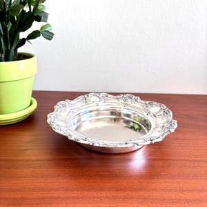May include: A round silver-colored metal dish featuring a smooth, reflective center basin and a wide, ornate rim decorated with intricate embossed floral and scroll patterns. The dish has a scalloped edge and a polished, metallic finish throughout.