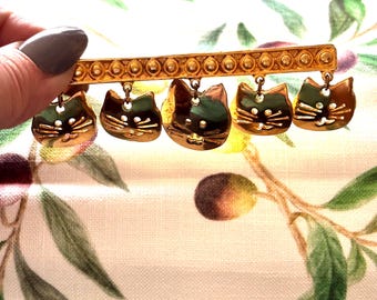 Op de afbeelding: Een goudkleurige broche met vijf hangende kattengezichten. De katten hebben gedetailleerde kenmerken en zijn opgehangen aan een rechthoekige staaf met een gestructureerd ontwerp. De broche wordt tegen een achtergrond van bedrukte stof gehouden.
