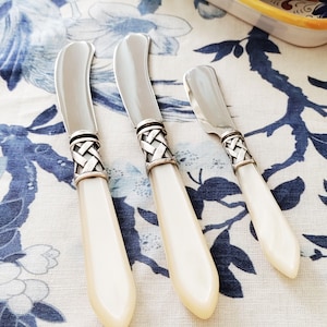May include: Three butter knives with silver blades and woven silver collars. The handles are a creamy off-white color. The knives are arranged on a blue and white floral patterned tablecloth.