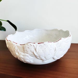 May include: A white ceramic bowl featuring a raised, embossed artichoke pattern around the exterior. The bowl has a scalloped, wavy rim with a thin, hand-painted reddish-brown line accentuating the edge of the ceramic structure.