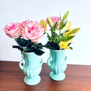 Puede incluir: Dos jarrones de cerámica azul claro a juego con asas ornamentadas y base acanalada. Un jarrón contiene un ramo de rosas rosas y blancas con hojas verdes, mientras que el otro contiene lirios amarillos y capullos de rosa con tallos verdes.