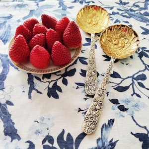 Könnte beinhalten: Ein Teller mit frischen, reifen Erdbeeren liegt neben zwei verzierten goldenen Servierlöffeln mit floralen Griffen. Die Löffel und der Teller befinden sich auf einer blau-weiß geblümten Tischdecke, was eine charmante, elegante Präsentation ergibt.
