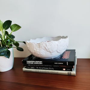 May include: A white ceramic bowl with a scalloped rim and embossed floral design. The bowl sits atop a stack of books with titles including "Twentieth Century Costume Jewellery". A potted plant is to the left.