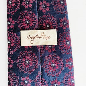 May include: A close-up of a navy blue tie with a repeating floral pattern in burgundy and white. The tie has a label that reads "Angelo Lica" in a cursive font.