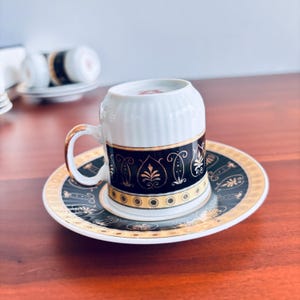 May include: A white porcelain coffee cup and saucer set featuring a black band with gold filigree patterns and a gold-trimmed handle. The cup is placed upside down on the saucer, which displays a matching black and gold decorative border design.