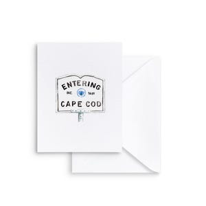May include: A white greeting card with a watercolor illustration of a sign that reads "Entering Cape Cod." The sign is in a rectangular shape with a blue emblem and the text "Inc. 1639." A white envelope is behind the card.