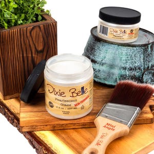 May include: An assortment of craft supplies, including Dixie Belle Pearlescent Glaze in a jar, a paintbrush, and a wooden planter with greenery. The glaze jar is open, and the brush has a wooden handle. The items are on a wooden surface.