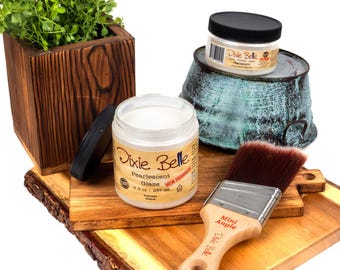 May include: An assortment of craft supplies, including Dixie Belle Pearlescent Glaze in a jar, a paintbrush, and a wooden planter with greenery. The glaze jar is open, and the brush has a wooden handle. The items are on a wooden surface.