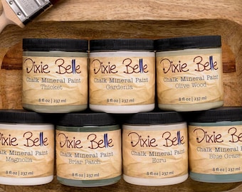 May include: Eight 8-ounce jars of Dixie Belle Chalk Mineral Paint in various colors, including Gardenia, Thicket, Olive Wood, Magnolia, Briar Patch, Ecru, and Blue Grass. The jars are arranged in two rows on a wooden surface.