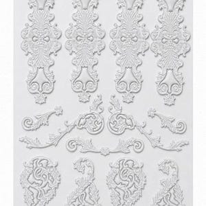 May include: A white silicone mold featuring various raised relief designs, including four elongated vertical scrollwork pieces, three horizontal ornate flourishes, and four teardrop-shaped decorative elements with intricate leaf and swirl patterns.