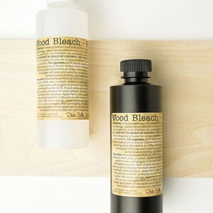May include: Two bottles of wood bleach are displayed against a light wood background. One bottle is white with a white cap, and the other is black with a black cap. Both bottles have labels with product information.