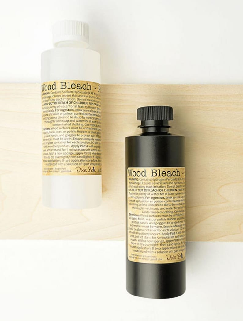 May include: Two bottles of wood bleach are displayed against a light wood background. One bottle is white with a white cap, and the other is black with a black cap. Both bottles have labels with product information.