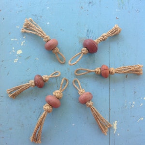 Op de afbeelding: Vijf handgemaakte rustieke ornamenten. Elk ornament heeft een roodbruine houten kraal, geknoopt jute touw en een kwast. De ornamenten zijn in een stervorm gerangschikt tegen een lichtblauwe, verweerde achtergrond.