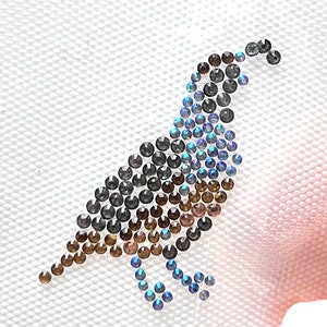 May include: A close-up of a small, brown and blue bird made of rhinestones. The bird is sitting on a white canvas background.