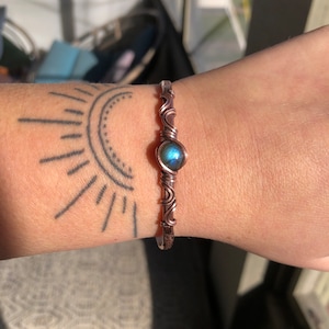 May include: Copper wire bracelet with a central, round, iridescent blue-green stone. The bracelet features intricate wire wrapping and a simple, open cuff design. The metal has a warm, reddish-brown hue.