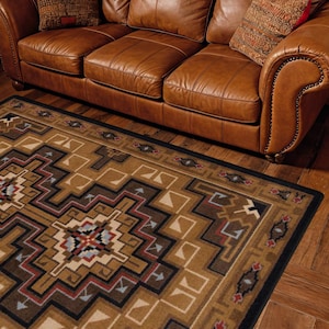 May include: A tan leather sofa with rolled arms and nailhead trim, featuring three seat cushions and three back cushions. Two patterned throw pillows are placed on the sofa. A rectangular rug with a geometric pattern in brown, tan, and blue.