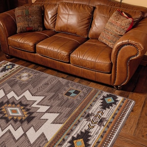May include: A three-cushion leather sofa in a warm tan color with rolled arms and nailhead trim, featuring two patterned throw pillows. Below the sofa is a rectangular area rug with a geometric pattern in shades of gray, brown, and teal.