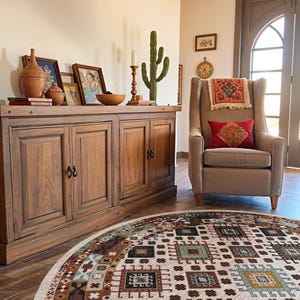 May include: A round area rug featuring a cream background with a repeating geometric pattern in shades of brown, teal, and black. The rug is positioned in front of a wooden sideboard and a beige upholstered armchair with a red accent pillow.