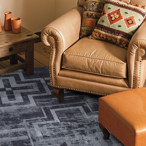 May include: A rectangular area rug featuring a dark blue and grey geometric pattern with intersecting lines and blocks. The rug is positioned on a wooden floor beneath a tan leather armchair, a matching leather ottoman, and a wooden side table.