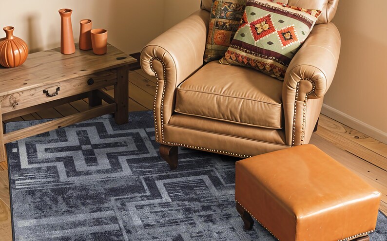May include: A rectangular area rug featuring a dark blue and grey geometric pattern with intersecting lines and blocks. The rug is positioned on a wooden floor beneath a tan leather armchair, a matching leather ottoman, and a wooden side table.