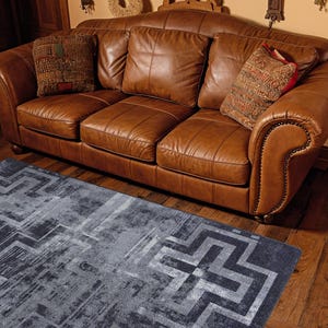 May include: A three-seat brown leather sofa with rolled arms and nailhead trim, featuring three back cushions and three seat cushions. Two decorative throw pillows with textured patterns are placed on the sofa. A grey rug with a geometric pattern.