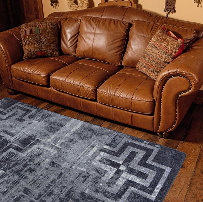 May include: A three-seat brown leather sofa with rolled arms and nailhead trim, featuring three back cushions and three seat cushions. Two decorative throw pillows with textured patterns are placed on the sofa. A grey rug with a geometric pattern.