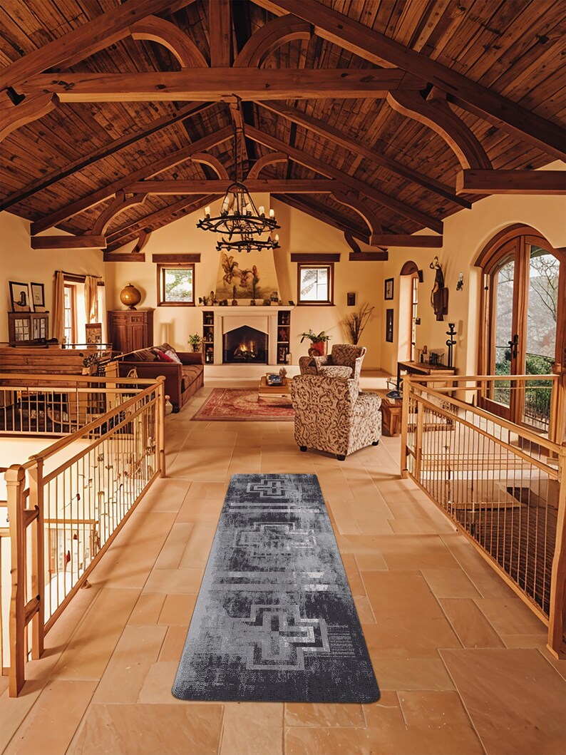 May include: A long, rectangular runner rug featuring a distressed, charcoal grey and light grey colour palette with a repeating geometric cross pattern. The rug is positioned centrally on a light-coloured stone floor in a large, open-plan room.