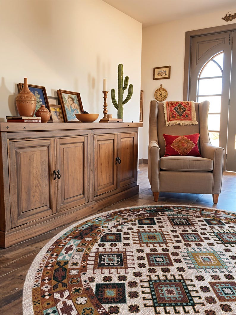May include: A round area rug featuring a cream background with a repeating geometric pattern in shades of brown, teal, and black. The rug is positioned in front of a wooden sideboard and a beige upholstered armchair with a red accent pillow.