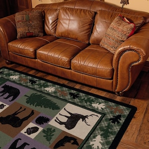 May include: A three-cushion tan leather sofa with nailhead trim and two patterned throw pillows, positioned above a rectangular area rug featuring a green plaid border and panels depicting black silhouettes of bears, deer, and fish.