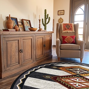 May include: A round area rug featuring a geometric pattern in black, white, rust orange, and olive green. The design includes a central diamond motif with intricate lines and angular shapes, surrounded by a wide black border with a white zigzag edge.