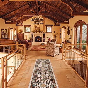 May include: A long rectangular runner rug featuring a cream-colored base with a repeating geometric pattern in shades of teal, orange, brown, and black. The rug is positioned on a light-colored stone floor in a large room with wooden beams.