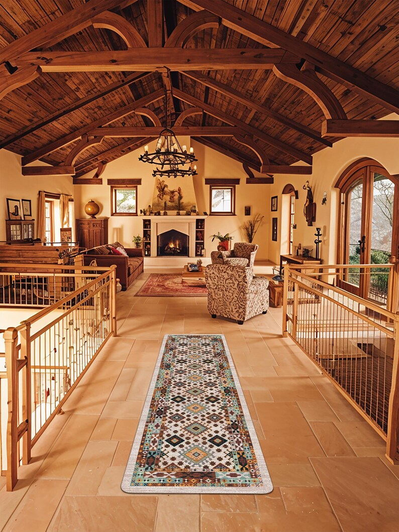 May include: A long rectangular runner rug featuring a cream-colored base with a repeating geometric pattern in shades of teal, orange, brown, and black. The rug is positioned on a light-colored stone floor in a large room with wooden beams.