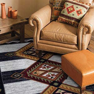 May include: A rectangular area rug featuring a geometric pattern with black, gray, and brown diamond shapes, accented by white zigzag borders. The rug is positioned on a wooden floor beneath a tan leather armchair, a matching ottoman, and a wooden table.