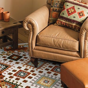 May include: A tan leather armchair with nailhead trim, a matching leather ottoman, and a patterned area rug featuring geometric shapes in orange, blue, and white. A wooden side table holds four terracotta-colored vases of varying heights.