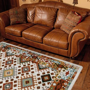 May include: A three-seat brown leather sofa with rolled arms and nailhead trim, featuring three back cushions and three seat cushions. Two patterned throw pillows are placed on the sofa, which sits above a white rug with geometric patterns.