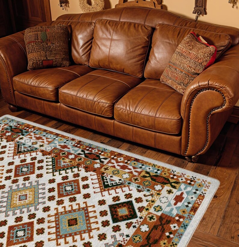 May include: A three-seat brown leather sofa with rolled arms and nailhead trim, featuring three back cushions and three seat cushions. Two patterned throw pillows are placed on the sofa, which sits above a white rug with geometric patterns.