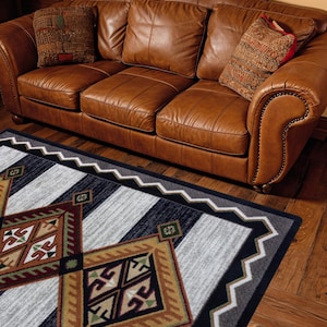 May include: A rectangular area rug featuring a geometric pattern with large diamonds in shades of tan, brown, black, and white. The rug has a zigzag border and is positioned in front of a brown leather sofa with three cushions and two patterned pillows.