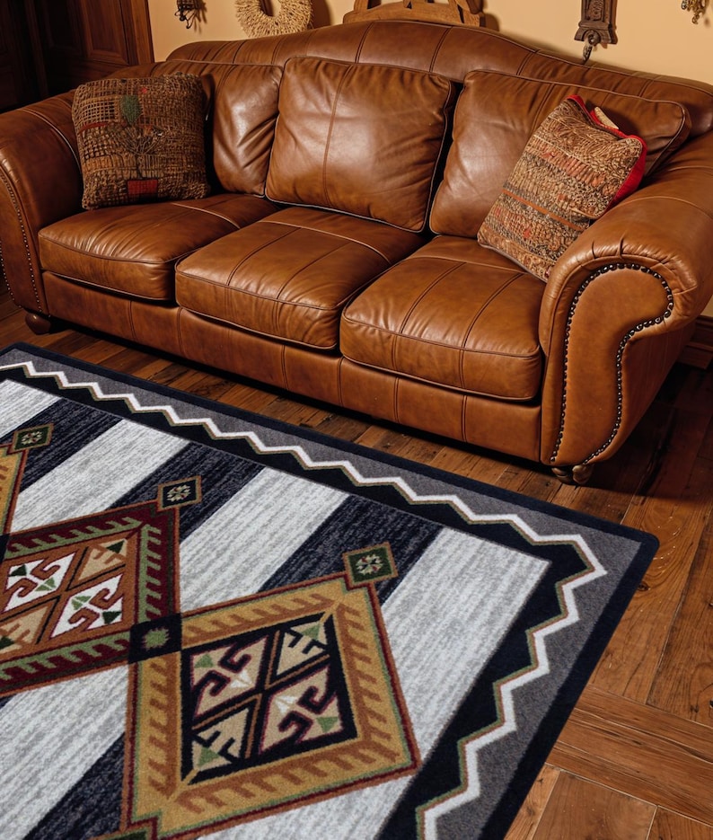 May include: A rectangular area rug featuring a geometric pattern with large diamonds in shades of tan, brown, black, and white. The rug has a zigzag border and is positioned in front of a brown leather sofa with three cushions and two patterned pillows.