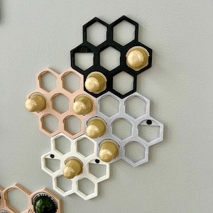 May include: Honeycomb-shaped coffee pod holders in black, white, pink, and silver, mounted on a gray wall. Each holder contains gold-colored coffee pods. The holders are arranged in a cluster, showcasing a modern and organized coffee storage solution.