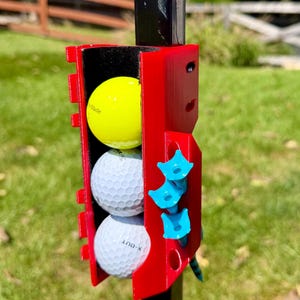 May include: A red plastic golf ball holder attached to a black pole, containing one yellow golf ball and two white golf balls stacked vertically. Three light blue golf tees are inserted into the side of the holder, which features a black interior lining.