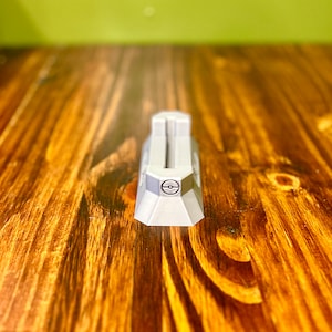 May include: A gray plastic Pokemon card holder with a Pokeball symbol on the top. The holder is sitting on a wooden surface.