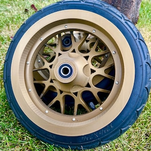May include: A gold-colored scooter wheel with a black tire. The wheel has a starburst design and is set against a backdrop of green grass and a tree trunk. The tire size is 12 1/2 X 2 1/4.