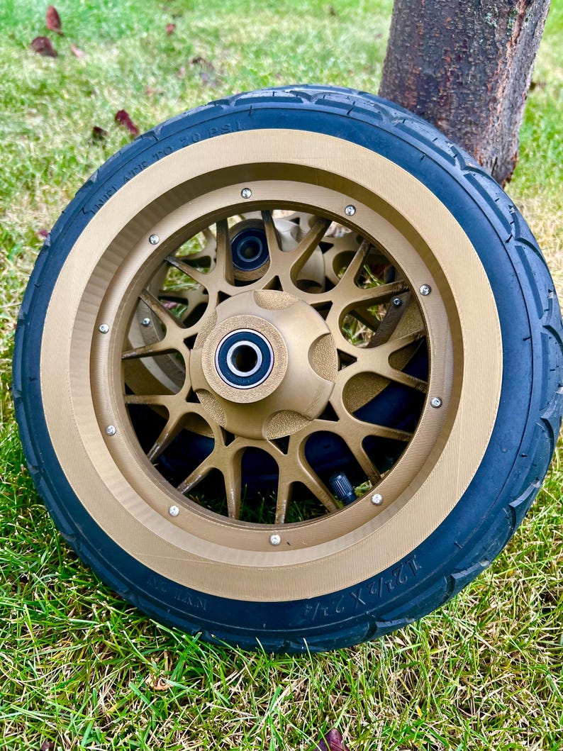 May include: A gold-colored scooter wheel with a black tire. The wheel has a starburst design and is set against a backdrop of green grass and a tree trunk. The tire size is 12 1/2 X 2 1/4.
