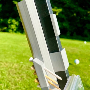 May include: Close-up of a golf tee holder attached to a black and gray post. The holder is light gray and holds several wooden golf tees. The background is a green lawn with a few white golf balls.