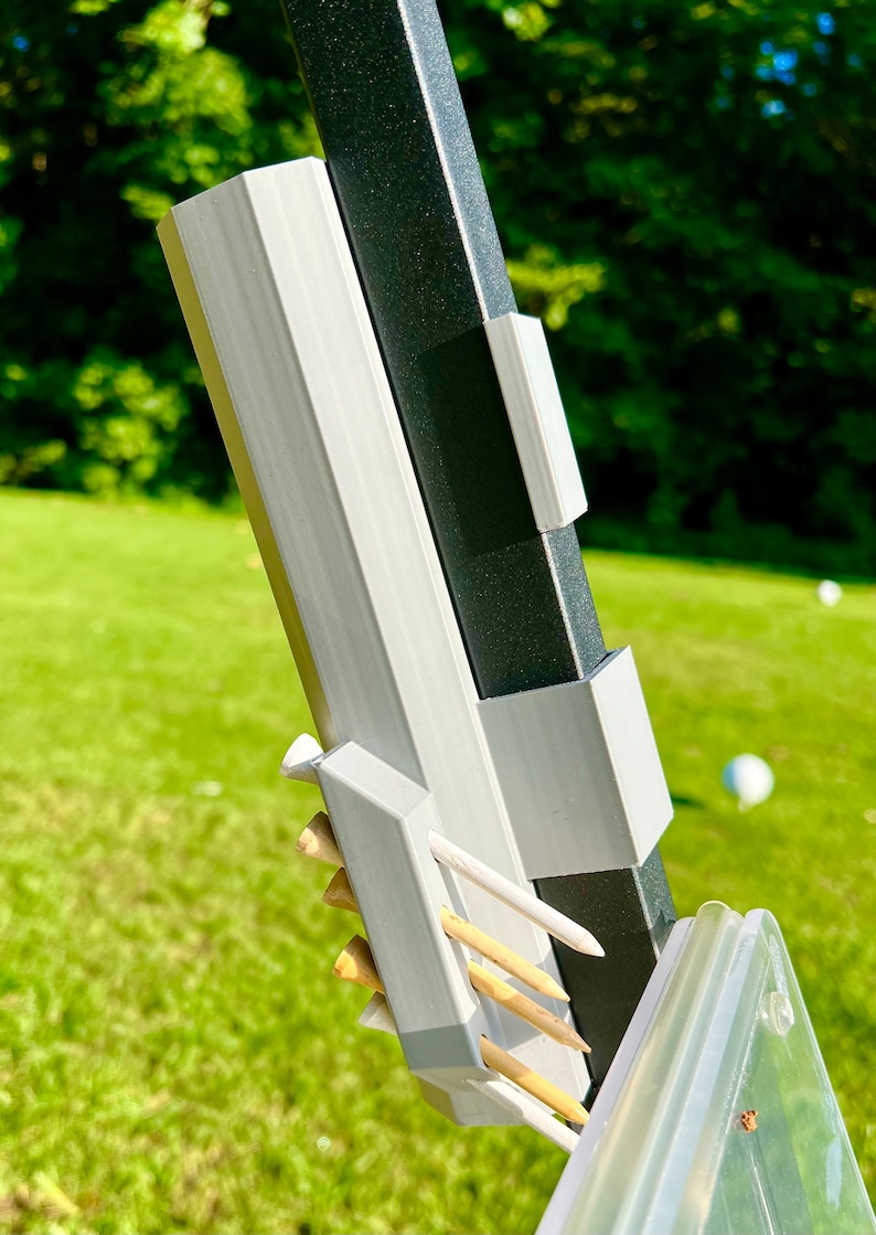 May include: Close-up of a golf tee holder attached to a black and gray post. The holder is light gray and holds several wooden golf tees. The background is a green lawn with a few white golf balls.