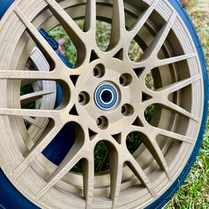May include: A close-up of a gold-colored car wheel with a black tire. The wheel has a multi-spoke design with several circular holes around the center. The tire has the text "20 PSI" printed on it. The wheel is on a grassy surface.