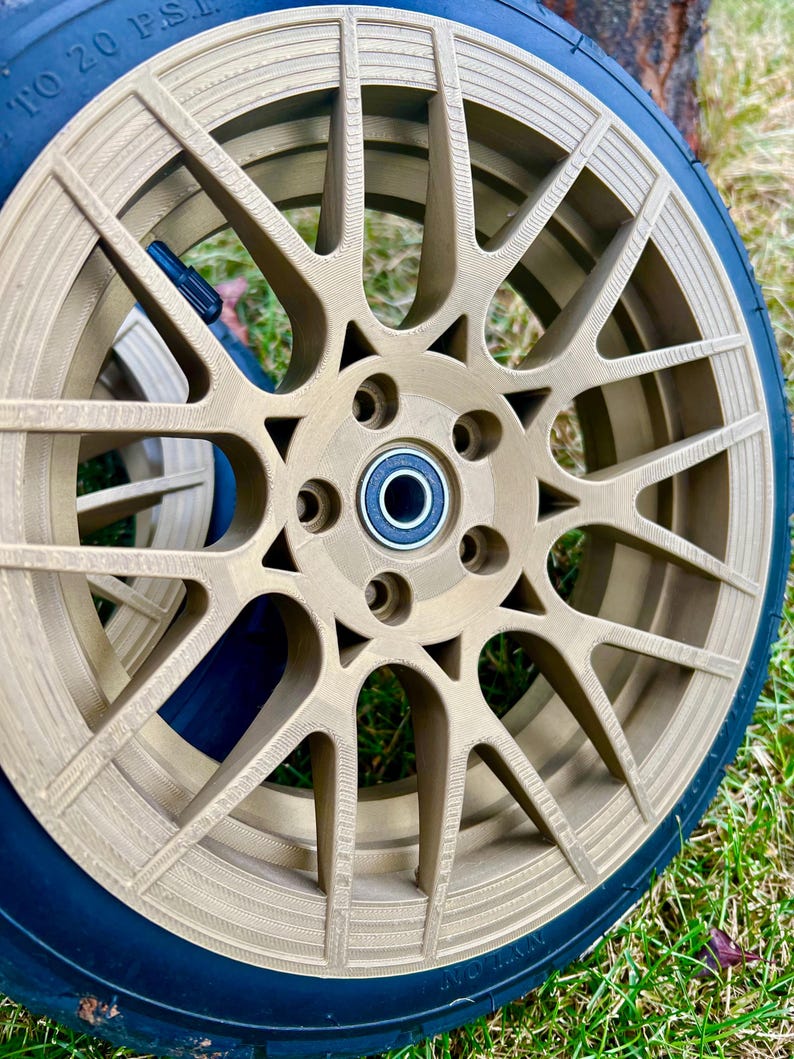 May include: A close-up of a gold-colored car wheel with a black tire. The wheel has a multi-spoke design with several circular holes around the center. The tire has the text "20 PSI" printed on it. The wheel is on a grassy surface.