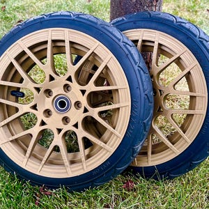 May include: Two gold-colored wheels with black tires are displayed on a grassy surface. The wheels have a complex spoke design and a central hub. The tires have a textured surface. The wheels are likely for a scooter or small vehicle.