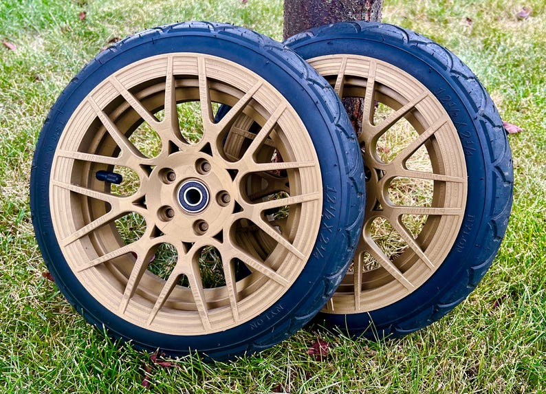 May include: Two gold-colored wheels with black tires are displayed on a grassy surface. The wheels have a complex spoke design and a central hub. The tires have a textured surface. The wheels are likely for a scooter or small vehicle.
